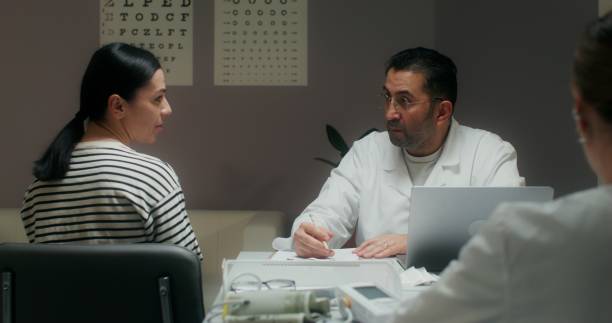 A female patient and a doctor talk sitting in a medical office. A man writes in a notebook, making an appointment for treatment